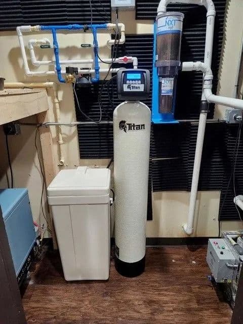 Water filtration system with a tall white tank, softener, and blue/white pipes in an enclosed space.
