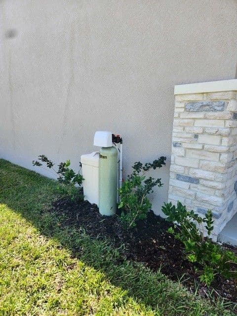 A Titan whole-home water treatment system next to a stucco wall and a decorative stone column. Bushes planted along the wall.