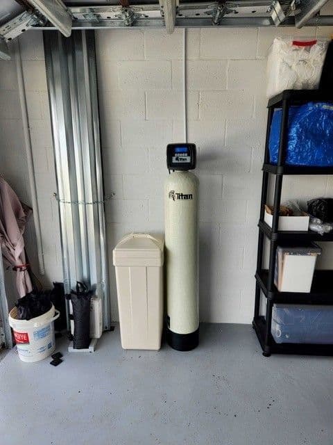 A Titan whole-home water treatment system, trash bin, and storage shelving in a garage. Beige and white tones.