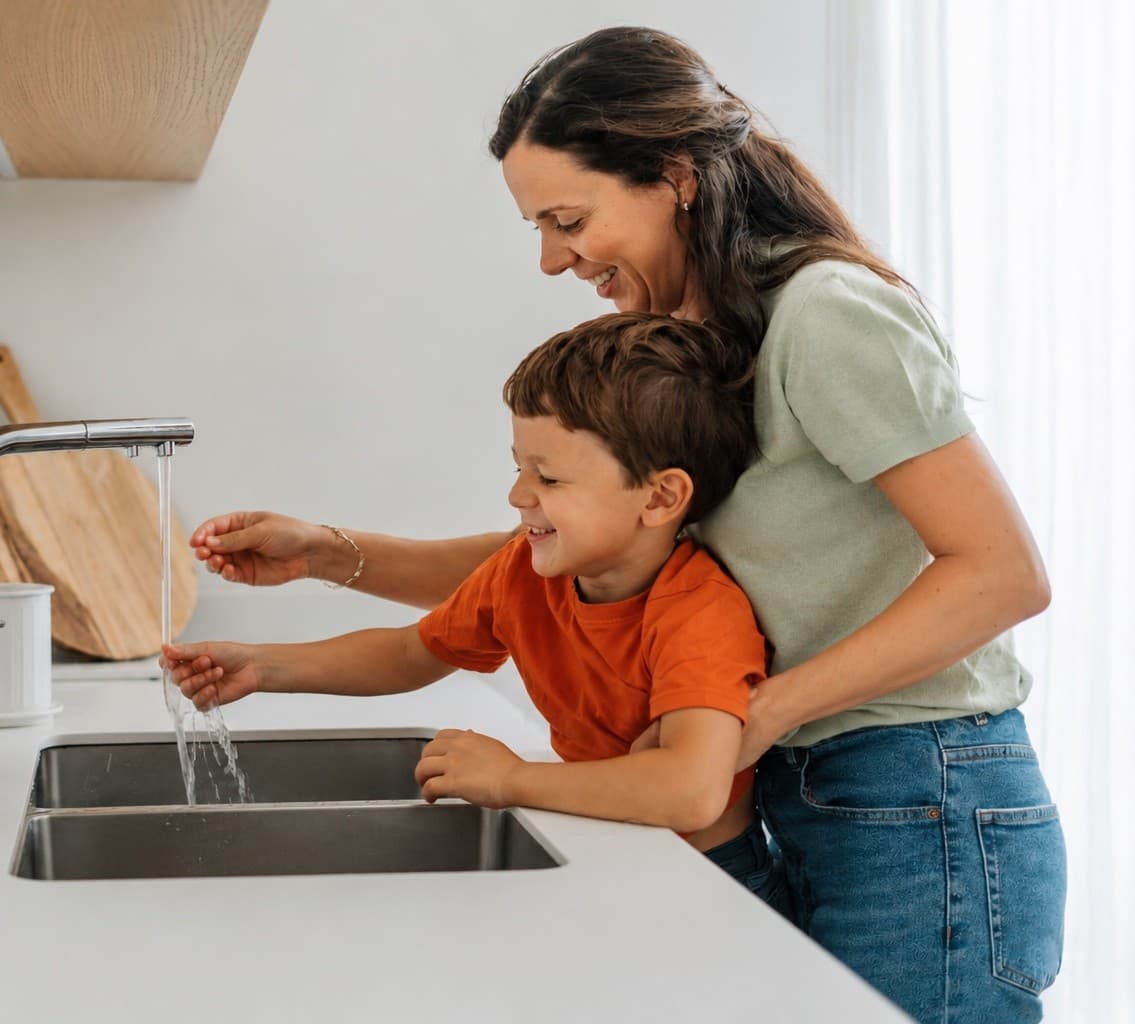 A mother and young son washing produce together at a bright kitchen sink